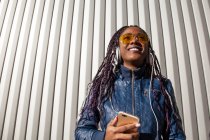 Von unten fröhliche junge Afroamerikanerin mit Afro-Zöpfen in blauer Jacke und stylischer Sonnenbrille, die Musik über Kopfhörer genießt, während sie im Sonnenlicht vor gestreifter Wand chillt — Stockfoto
