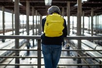 Вид ззаду невідомий чоловічий мандрівник з рюкзаком, що стоїть біля перил на переході над поїздами на залізничній станції — стокове фото