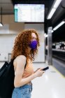 Vista lateral de mujer en máscara textil con teléfono celular y mochila mirando hacia otro lado en la plataforma del metro - foto de stock