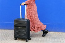 Side view of cropped unrecognizable female in long red dress standing with luggage on street against blue wall in daytime — Stock Photo