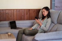 Молодая этническая женщина со стеклом кофе серфинг интернет на мобильном телефоне сидя на диване в домашней комнате рядом с ноутбуком — стоковое фото