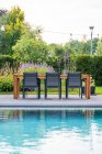 Обеденный стол со стулом и деревянными шезлонгами рядом с бассейном во дворе дорогой современной виллы в стиле минимализма в солнечный день — стоковое фото