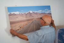 Vista posterior de hombre calvo senior en anteojos y camisa a cuadros colgando obras de arte de la carretera y las montañas en la pared en casa - foto de stock