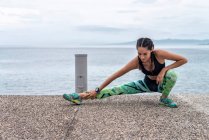 Focused female athlete doing side lunge exercise and stretching legs while warming up during training on embankment near sea — Stock Photo