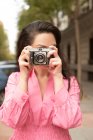 Joven mujer feliz con el pelo largo y castaño tomando fotos en la vieja cámara de fotos en la calle en la ciudad - foto de stock