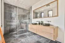Éviers en céramique sur placard en bois sous miroir suspendu au mur dans la salle de bain avec murs carrelés et unité de douche — Photo de stock