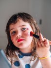 Criança com aplicador fazendo face com produtos cosméticos variados em casa olhando para a câmera — Fotografia de Stock