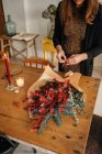 Сверху неузнаваемая женщина-флористка стоит за деревянным столом со свечами и устраивает рождественский букет в комнате — стоковое фото