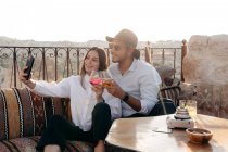 Позитивная молодая и стильная пара, сидящая на диване и звенящие стаканы коктейля, делая автопортрет на сотовом телефоне на террасе в Каппадокии, Турция — стоковое фото