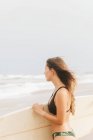 Ragionevole giovane atleta donna in costume da bagno con capelli volanti e tavola da surf guardando lontano sulla costa dell'oceano — Foto stock
