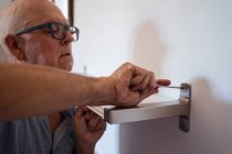 Внимательный пожилой мужчина в очках с ручной отвёрткой, прикручивающей полку к стене в комнате — стоковое фото