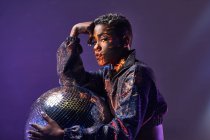 Seitenansicht der trendigen jungen Afroamerikanerin mit Make-up, die in die Kamera schaut, während sie die Glitzerkugel in violettem Licht hält — Stockfoto