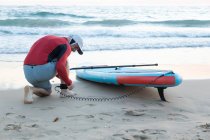 Visão traseira do surfista masculino em wetsuit colocando trela de tornozelo na placa SUP enquanto se prepara para remar surf na costa — Fotografia de Stock