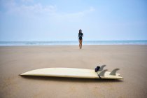 Ethnische Surferin im Neoprenanzug spaziert an der Küste gegen Surfbrett unter wolkenlosem blauen Himmel — Stockfoto