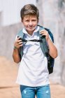 Zufriedenes Schulkind in zerrissenen Jeans und Gummischuhen blickt auf gefliestem Bürgersteig in sonniger Stadt in die Kamera — Stockfoto