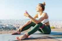 Young contemplative female athlete with bottle of water and resistance band touching forehead while looking away on urban rooftop — Stock Photo