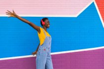 Mujer afroamericana despreocupada escuchando música en auriculares inalámbricos y brazos extendidos contra una pared brillante - foto de stock