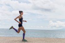 Vista lateral del corredor femenino en forma en el momento de saltar sobre el paseo marítimo durante el entrenamiento cardiovascular enérgico en verano - foto de stock