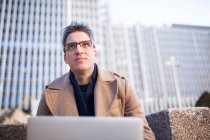 Niedriger Winkel des jungen Mannes in trendigem Outfit und Brille sitzt auf einer Bank und blättert im Netbook, während er an einem Projekt auf der Straße arbeitet — Stockfoto