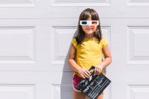 Linda chica feliz en ropa colorida casual sosteniendo clapperboard mientras que en gafas tridimensionales de pie sobre fondo blanco de la pared - foto de stock