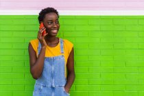 Contenido Mujer afroamericana de pie con la mano en el bolsillo cerca de la pared vívida y hablando por teléfono celular mientras mira hacia otro lado - foto de stock
