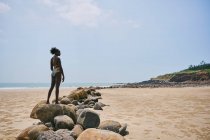 Side view of content young African American female in swimsuit standing on boulder while looking away — Stock Photo