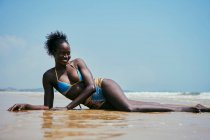 Joven y alegre hembra étnica en traje de baño con moño de pelo afro mirando hacia otro lado mientras yace en la costa del océano bajo el cielo azul - foto de stock