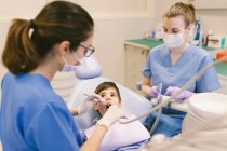Alto ángulo de dentista y asistente de tratamiento de dientes de niño durante el procedimiento en la clínica de odontología - foto de stock