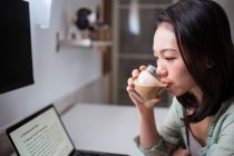 Вид збоку молодої етнічної жінки блогер за столом з нетбуком і кавою, дивлячись на камеру в домашній кімнаті — стокове фото