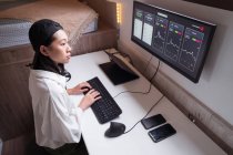 Vue latérale à angle élevé d'une femme asiatique concentrée travaillant sur ordinateur avec des graphiques montrant la dynamique des changements de valeur de la crypto-monnaie sur un lieu de travail pratique — Photo de stock
