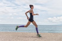 Vista lateral del corredor femenino en forma en el momento de saltar sobre el paseo marítimo durante el entrenamiento cardiovascular enérgico en verano - foto de stock