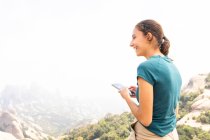 Vista lateral de mensajes de texto de turismo femenino positivo en el teléfono celular durante la excursión en las montañas de Montserrat en España - foto de stock