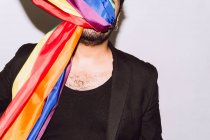 Homme homosexuel méconnaissable avec le visage enveloppé dans le drapeau arc-en-ciel symbole de la communauté LGBT sur fond blanc — Photo de stock