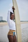 Vue latérale d'une athlète afro-américaine avec planche de surf admirant l'océan depuis un rivage sablonneux sous un ciel bleu nuageux — Photo de stock