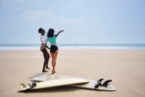 Surfeur méconnaissable pointant vers l'océan tout en interagissant avec la petite amie noire joyeuse contre longboard et planche de surf sur la côte sablonneuse — Photo de stock