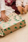 Récolter anonyme enfant ouverture boîte cadeau avec motif sapin sur le sol pendant les vacances du Nouvel An dans la maison — Photo de stock