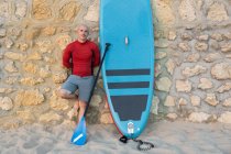 Surfeur masculin en combinaison et chapeau debout regardant la caméra avec pagaie et planche SUP tout en se préparant à surfer sur le bord de la mer appuyé sur un mur de pierre — Photo de stock