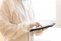 Médico masculino irreconocible cultivado en equipo de protección personal que toca la pantalla en la tableta durante la pandemia de COVID 19 en la clínica - foto de stock