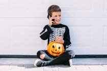 Полное тело веселого мальчика в костюме черного скелета с раскрашенным лицом и вырезанной тыквой на Хэллоуин, разговаривающей по мобильному телефону, сидя возле белой стены на улице — стоковое фото