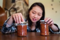 Young ethnic female with glass jars of delicious fig marmalade on table in house on blurred background — Stock Photo