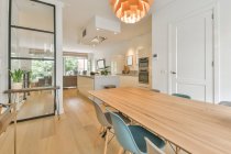 Современный интерьер кухни открытой планировки столовой с деревянным столом и пластиковыми стульями под креативной люстрой в просторной новой квартире — стоковое фото