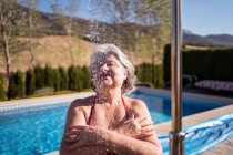 Allegro anziana femmina in bikini godendo spruzzi da doccia vicino piscina con acqua limpida trasparente — Foto stock