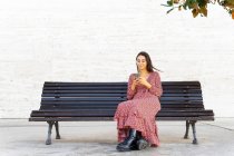 Corps complet de femme positive dans des vêtements élégants navigation smartphone et assis sur un banc en bois sur la rue en journée — Photo de stock