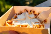 Cortada pessoa irreconhecível comendo waffles belgas saborosos com chantilly em caixa takeaway contra montagens em back lit — Fotografia de Stock