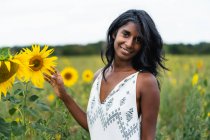 Mulher étnica adulta sincera olhando para a câmera no prado tocando flores florescentes no campo no fundo borrado — Fotografia de Stock