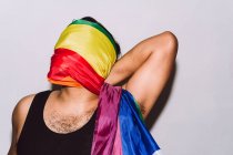 Maschio omosessuale irriconoscibile con volto avvolto nella bandiera arcobaleno simbolo della comunità LGBT su sfondo bianco — Foto stock