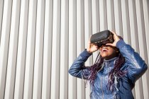Задоволена молода афроамериканська жінка в VR хедсеті розважається і грає віртуальну гру проти сірої смугасті стіни — стокове фото