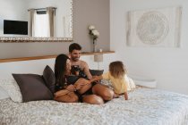 Bambino accarezzando pancia sorridente incinta mamma in lingerie sdraiata sul letto con marito in casa — Foto stock