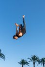 Vista lateral de ángulo bajo del atleta masculino activo haciendo backbend mientras que salta contra palmeras bajo cielo azul en la luz del sol - foto de stock