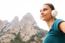 Jovem turista sonhadora olhando para longe desfrutando de música de fone de ouvido sem fio contra Montserrat e árvores na Espanha — Fotografia de Stock